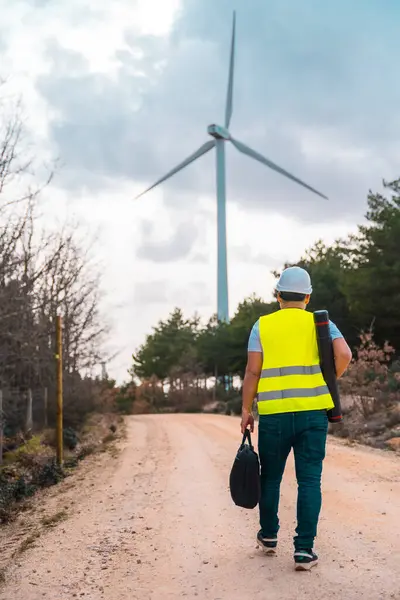 一个男性高加索裔成年工程师沿着绿色能源公园的小径走着 带着风力涡轮机的后视镜的垂直照片 图库图片