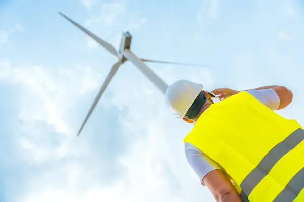 一位男性高加索成人工程师在一个绿色能源园区的风力涡轮机旁边用手机拍摄的低视角照片 免版税图库照片