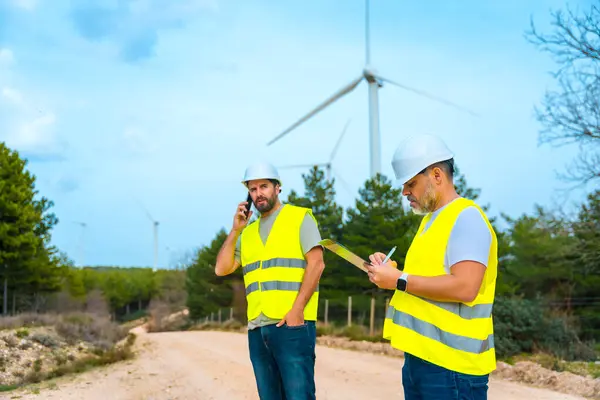 两名男性同事工程师在风力涡轮机旁边的一个绿色公园工作 免版税图库图片