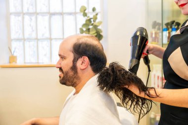 Hairdresser drying a washed capillary prosthesis before attaching it to the head of a man clipart