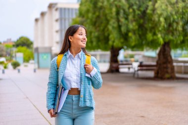 Mutlu Asyalı üniversite öğrencisi kampüste yürürken gülümsüyor ve yan bakıyor.