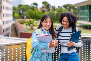 İki güzel, genç, gülümseyen çoklu etnik öğrenci üniversite kampüsünün dışında duran notları okuyorlar.