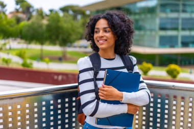 Latin kız öğrenci kampüsün dışında elinde dosya ve sırt çantası tutarak gülümsüyor.