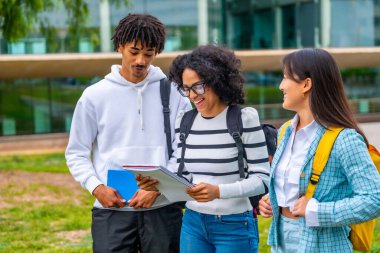 Üniversitenin dışında duran notları okurken gülümseyen üç çok ırklı öğrenci.