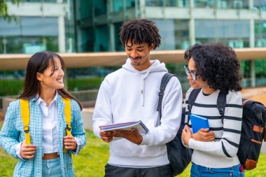 Modern ve yeşil bir kampüste, üniversitenin dışında duran çok ırklı üniversite öğrencileri birlikte notlar okuyorlar.