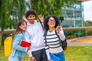 Üç çok ırklı öğrenci kampüsün dışında takılabilir bir kamerayla bir video kaydederken el sallıyor.