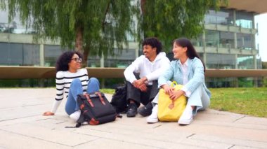 Üniversitede rahatça oturup sohbet eden üç gülümseyen çok ırklı öğrencinin tam boy fotoğrafı.
