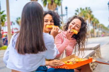 Mutlu çok ırklı kadınlar pizzalarını paylaşıyor. Yaz günü sokakta oturuyorlar.