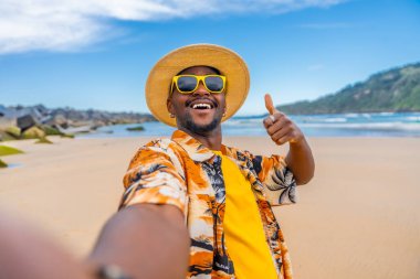 Yazlık kıyafetleri olan havalı bir adam baş parmağıyla sahilde selfie çekiyor.