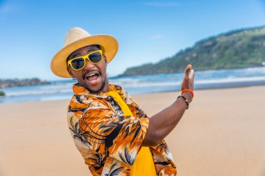 Yazlık elbiseli, güneş şapkalı ve güneş gözlüklü mutlu afro adam kumlu bir sahili işaret ediyor.