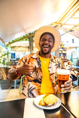 Terasta bira ve sandviçin keyfini çıkaran mutlu Afro-Amerikan turistin dikey fotoğrafı.