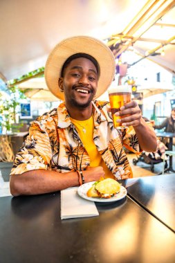 Terasta bira ve sandviçin keyfini çıkaran mutlu esmer genç bir erkek turistin dikey fotoğrafı.