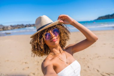 Personal perspective of a beauty young latin woman holding a sun hat attached at her head while taking selfie a beach