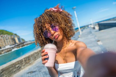Personal perspective of a cool latin young woman with curly hair and sunglasses drinking milkshake taking a selfie on the promenade