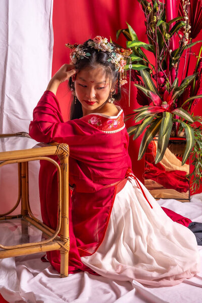 A young woman wearing a red and white dress sits on a wooden chair. She is wearing a flower headband and has red lipstick on. The image has a traditional and elegant feel to it