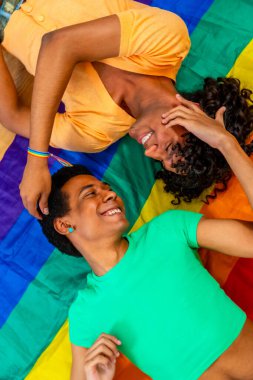 Çok ırklı eşcinsel bir çiftin LGBT gökkuşağı bayrağı altında yan yana yattığını gösteren dikey bir fotoğraf.
