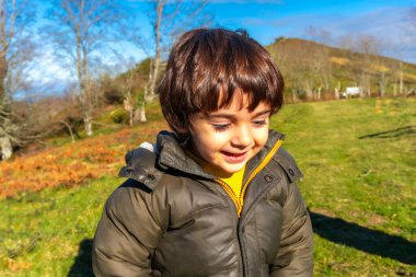 Yeşil ceketli ve gülümseyen bir çocuk tarlada duruyor. Mutluluk ve sıcaklık kavramı, çocuk dışarıda eğleniyor gibi görünüyor.