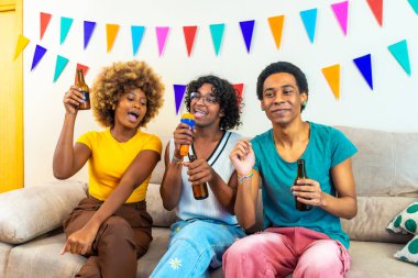 Karaoke sırasında şarkı söyleyen ve bira içen çeşitli arkadaşlar kanepede oturmuş duvarında çok renkli çelenkler var.