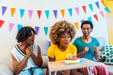 Doğum günü pastasına mum üfleyen kıvırcık saçlı Afrikalı genç bir kadın evde çok ırklı arkadaşlarıyla kanepede oturuyor.