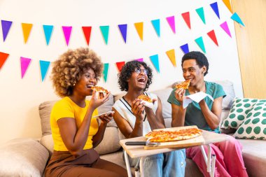 Farklı arkadaşlar bir dilim pizza yiyorlar. Kanepede oturmuş, evde kutlama yapıyorlar.