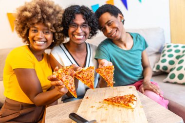 Çok ırklı arkadaşlar kameraya gülümseyen bir dilim pizza gösteriyorlar. Kanepede birlikte oturuyorlar.