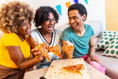 Üç farklı arkadaş kanepede oturmuş pizza paylaşıyor. Gülüyor ve evde eğleniyorlar.