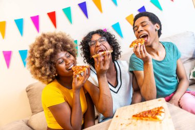 Çok ırklı arkadaşlar pizza yiyorlar. Evdeki kanepede birlikte oturarak eğleniyorlar.