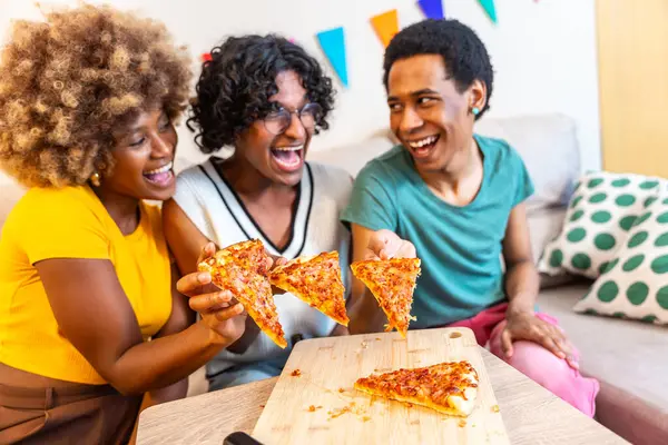 Üç farklı arkadaş kanepede oturmuş pizza paylaşıyor. Gülüyor ve evde eğleniyorlar.