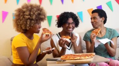 Farklı arkadaşlar bir dilim pizza yiyorlar. Kanepede oturmuş, evde kutlama yapıyorlar.