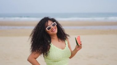 Bir kadın sahilde karpuz tutuyor. Gülümsüyor ve güneş gözlüğü takıyor. Kumsal güzel ve rahatlatıcı bir yer, ve kadın eğleniyor gibi görünüyor.