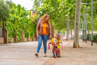 Sarışın anne ve kız bir parkın yanında bisiklet sürüyor ve yürüyor.