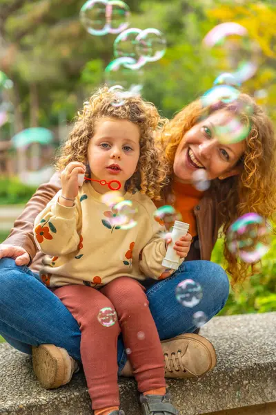 Parkta oturmuş baloncukları üfleyen tatlı sarışın bir kız ve annenin dikey portresi.