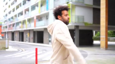 Şehirde dans eden siyah etnik adam beyaz kapüşonlu bir kapüşonlu giyen bir adam bir binanın önünde caddeden aşağı yürüyor. Sahne sakin ve huzurlu, adamın gökyüzüne baktığı