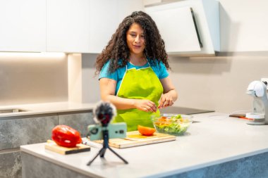 Latin genç bir kadın evdeki sağlıklı yiyeceklerin çevrimiçi videosunu çekiyor. Mutfakta cep telefonu ve tripodla kaydediyor.