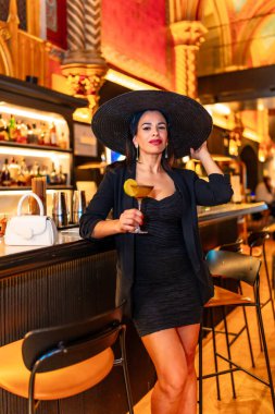 Lüks bir barda zarif şapkalı ve şehvetli elbisesi olan bir kadının dikey fotoğrafı.