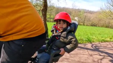 Bir adam çocuk koltuğunda bir çocukla bisiklete biniyor. Çocuk kask takıyor ve gülümsüyor.