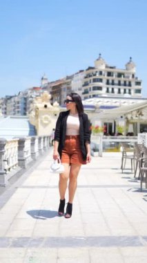 Modern bir kadının deniz kenarında yürüdüğü dikey bir video. Moda konsepti