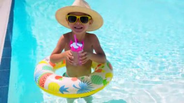 Güneş şapkalı, güneş gözlüklü, kauçuk bir halkanın içinde duran bir bardak suyla havuzda gezen beyaz bir çocuk.