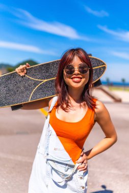 Omuzlarında paten taşıyan, yaz kıyafetleri ve güneş gözlükleriyle genç bir kadının dikey portresi.
