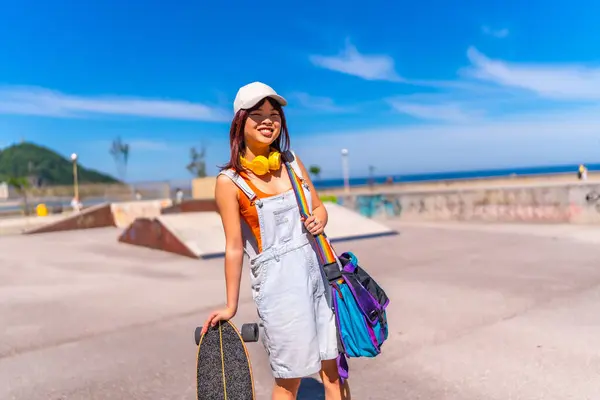 stock image Cute chinese woman in denim clothes and cap standing in a skateboard park in a sunny day holding an skate