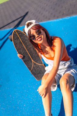 Modern elbiseli ve güneş gözlüklü mutlu bir patencinin dikey portresi mavi renkli parka oturmuş kameraya gülümsüyor.