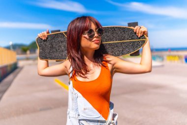 Kentli kendinden emin Asyalı bir kadının portresi kaykayı omuzlarında tutuyor yazın güneşin altındaki bir kaykay parkında duruyor.