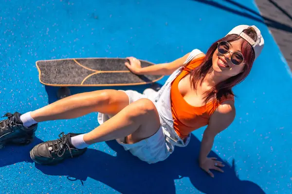 Güneş gözlüklü ve şapkalı havalı bir kadının renkli bir parkta kaykayıyla oturuşunun en güzel görüntüsü.