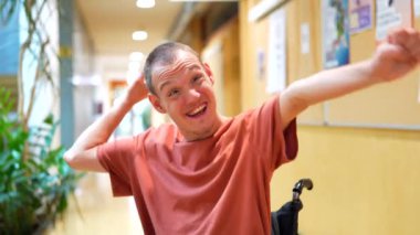 Tekerlekli sandalyede oturan engelli bir adam iş birliği koridorunda kameraya gülümsüyor.