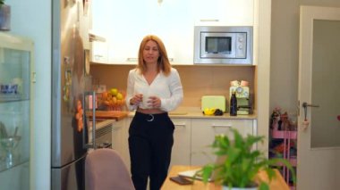 Beyaz tenli, sarışın, yetişkin bir kadının ev ofisinde çalışırken kahve hazırlarken geniş açılı görüntüsü.