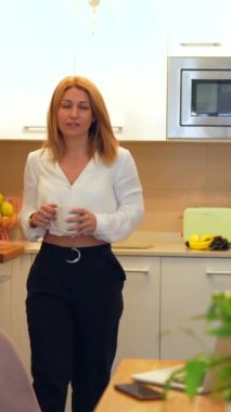 Sarışın, beyaz tenli, gülümseyen bir iş kadınının tele-işe giderken mutfakta dizüstü bilgisayar kullanarak telefonu açtığı dikey bir video.