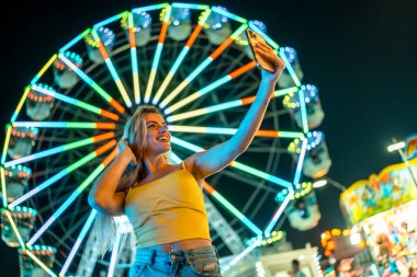 Sarışın bir kadının gece lunaparkta selfie çekerken düşük açılı görüntüsü.