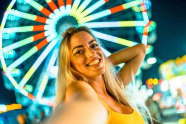 Şahsi bakış açısına göre güzel, mutlu bir kadın eğlence parkında selfie çekiyor ve arka planda neon ışıklarla aydınlatılan büyük bir tekerlek var.