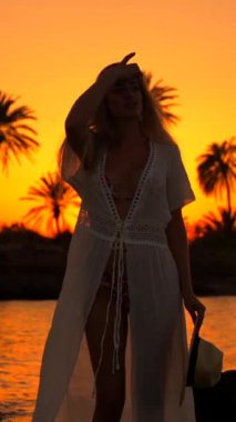 Tropikal plajda gün batımının keyfini çıkaran beyaz bir kadının dikey fotoğrafı.
