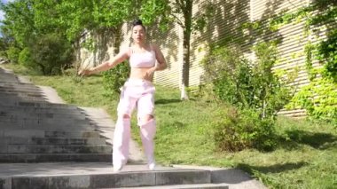 Pembeli bir kadın merdivenlerde dans ediyor. Pembe bluz ve pembe pantolon giyiyor.